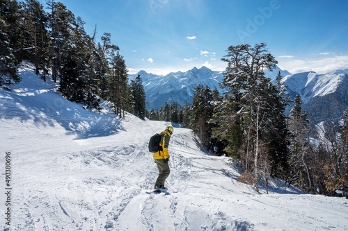 Beautiful landscape of the Arkhyz ski resort with mountains, snow, forest and man snowboarder in yellow jacket and backpack on a sunny winter day. Caucasus Mountains, Russia