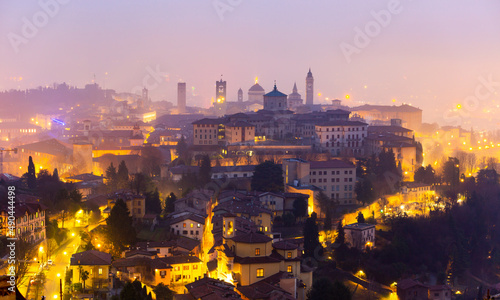 Picturesque night view of old fortified Upper City of Bergamo on hill with brightly lit streets and historic buildings in winter haze, Italy