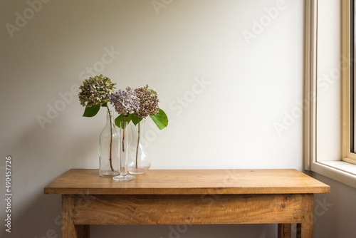 Dried hydrangea flowers in glass vases on oak side table next to window against beige wall (selective focus)