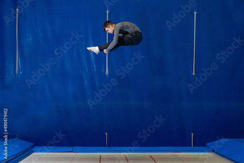 Young man doing an acrobatic figure on elastic bed, in blue background