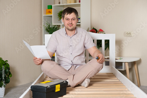 The furniture assembler assembles a children's white wooden bed, studies the assembly instructions