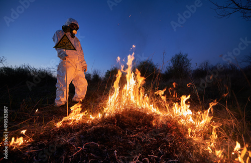 Fireman ecologist extinguishing fire in field at night. Man in suit and gas mask near burning grass, holding yellow triangle with skull and crossbones warning sign. Natural disaster concept.