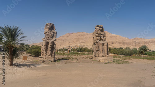 Giant statues of the colossi of Memnon against the blue sky and sand dune. Huge sculptures of seated pharaohs stand in the valley. Green vegetation in the distance. Egypt