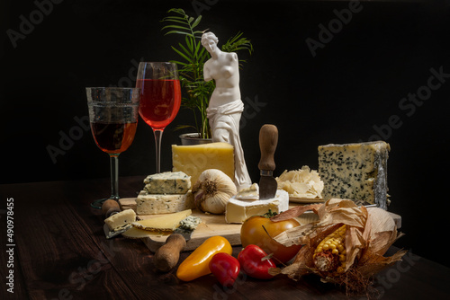 Still life with a wooden board full of delicious cheeses, tomatoes, strong garlic, sweet potatoes, red and green peppers, some glasses of rosé wine and lambrusco and a Greek female figure