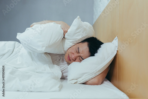 A man at home tries to fall asleep, noisy neighbors interfere with sleep, an Asian closes his ears with pillows, tired after work