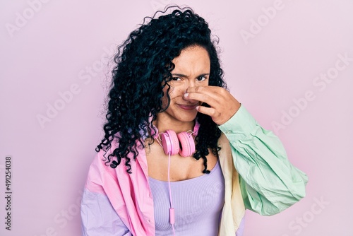 Young hispanic woman with curly hair wearing gym clothes and using headphones smelling something stinky and disgusting, intolerable smell, holding breath with fingers on nose. bad smell