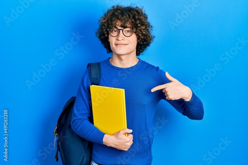 Handsome young man wearing student backpack and holding book pointing finger to one self smiling happy and proud