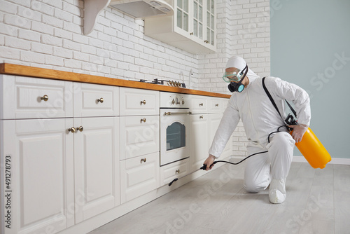 Pest control home service guy fighting parasites in the house. Exterminator in mask, goggles and PPE suit spraying poisonous gas or liquid from sprayer bottle on floor and cupboard in kitchen interior