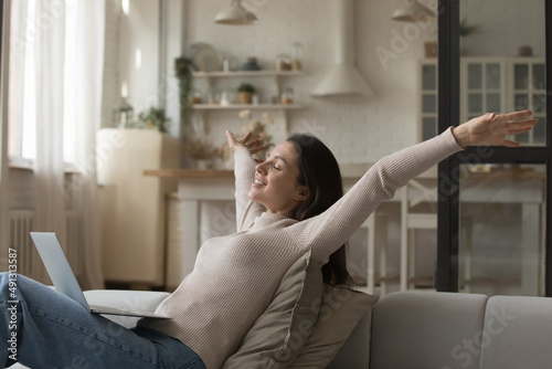 Happy young woman relaxing alone on cozy sofa put wireless computer on laps, raising her arms, female smiling with eyes closed enjoy carefree untroubled weekend, accomplish online task feeling free