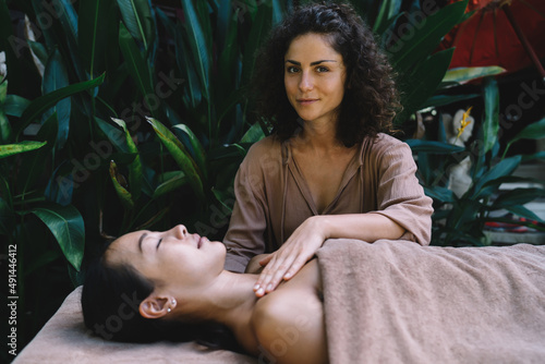 Portrait of Caucasian woman doing professional Thai massage to Asian female client enjoying technique of procedure, cosmetologist doing rejuvenation for stretch skin and looking at camera during seans