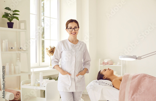 Portrait of friendly female woman beautician, aesthetic nurse or masseuse at her workplace. Smiling female beauty salon worker standing next to her client lying on couch in bright office.