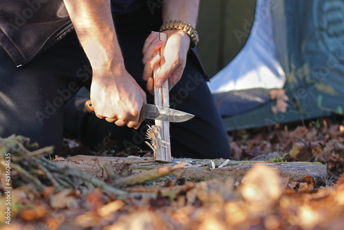 Close up of the hands of a man carving off timber to lit a fire, camp site and tent on the background. Hand holding knife cutting a wooden stick. Bushcraft and outdoors survival activities concept.