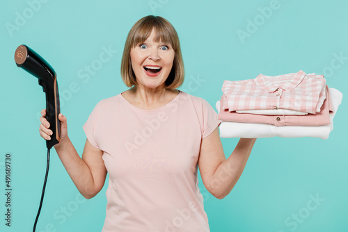 Elderly amazed shocked housewife woman 50s in pink t-shirt doing housework hold pile of clothes steamer isolated on plain pastel light blue background studio Housekeeping cleaning tidying up concept.