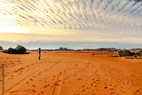 Unrecognizable tuareg man standing in the sahara desert. Colorful sand, Rocky Mountains, sky with sunlight diffusing in stratus clouds.