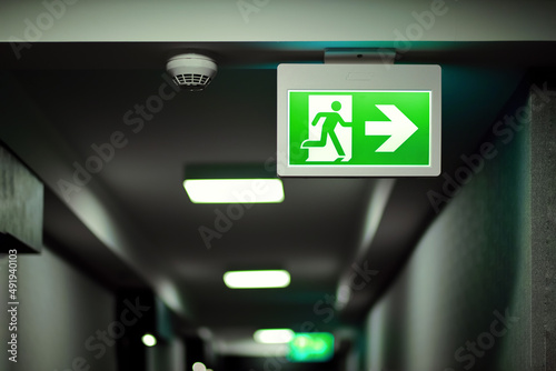 Green fire exit sign on hallway aside of smoke alarm device. Fire prevention and building safety background.