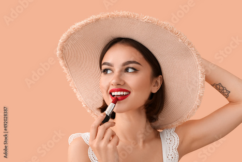 Happy woman in hat adjusting red lipstick on beige background