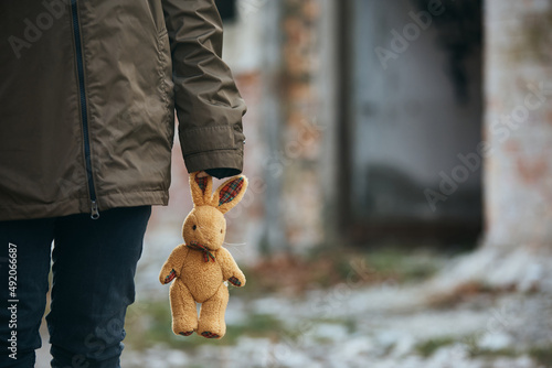 Unrecognizable refugee with plush bunny on war damaged city street.