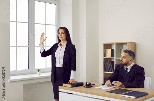 Young woman takes an oath before giving a sworn testimony during the court trial. Crime witness standing by the judge in the courtroom swears on the Holy Bible that she will tell nothing but the truth