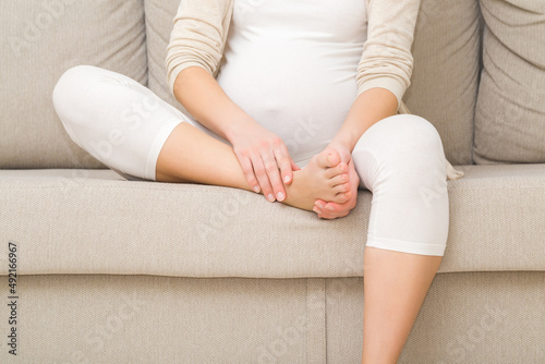 Young adult pregnant woman sitting on sofa and massaging her painful foot. Body health problem in pregnancy time. Closeup. Front view.