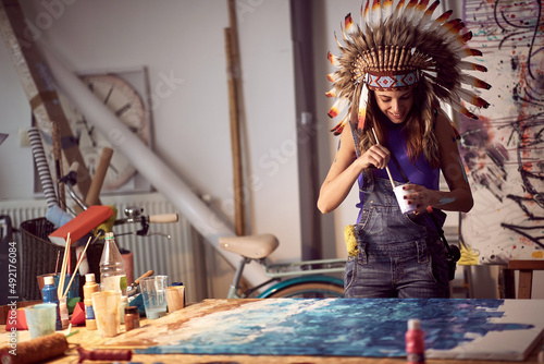 A young female artist with a war bonnet in the studio is mixing and preparing the paint for a new painting. Art, painting, studio
