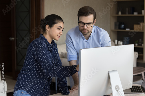 Millennial Indian woman stand by workplace desk discuss work on project on pc screen with focused man colleague teammate. Young businesswoman assist male partner give advice professional consultation