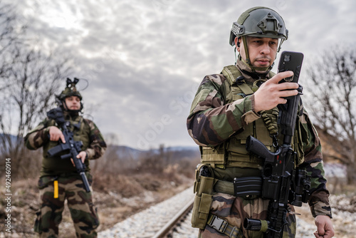 Two soldiers dogs of war mercenaries men in uniform armed service rifles standing while securing the railroad in combat zone in war soldier use mobile phone while patrolling area during mission