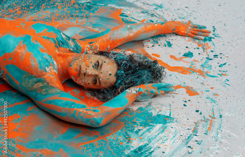 Head and upper body of expressive sexy naked woman lying elegant on the floor in turquoise blue orange color abstractly painted torso bodypainting woman on the splashed ground