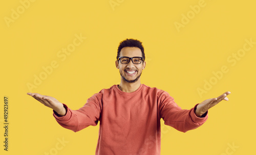 You did it. Congratulations. Happy cheerful positive young ethnic man in eyeglasses standing against yellow color background looking at camera, smiling and spreading his arms wide open for a warm hug