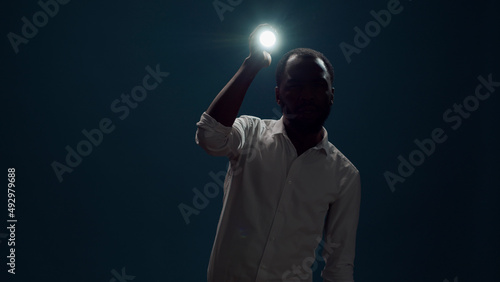 Young person sitting in the dark and using flashlight to search around, looking for evidence. Male detective taking risk and holding lantern to investigate and examine creepy sound.