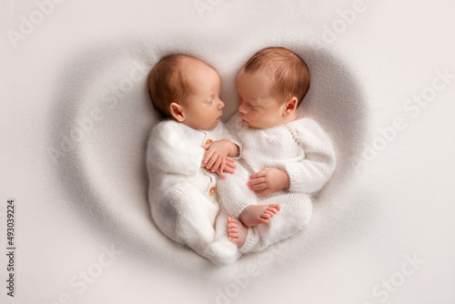 Tiny newborn twin boys in white bodysuits on a white background. Newborn twins sleep next to their brother on the background of the heart. Two newborn twin boys hugging each other.