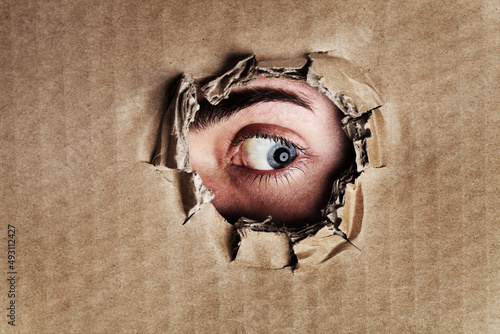 What is on the other side. Closeup portrait of an eye looking through a ripped hole in a piece of cardboard.