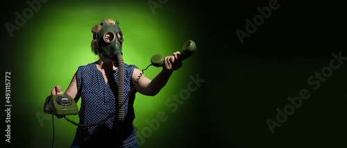 A woman in a gas mask passes the receiver of a retro phone on a dark dramatic background with copy space, hard light.