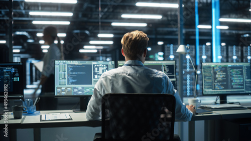 Back View of Skilled Focused System Administrator or Consultant Checks and Saves Backup Data in Dark Evening Room Workplace Station Indoors. Network Technologies Concept