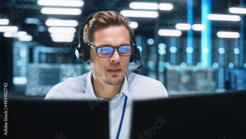 Portrait of a Young Caucasian Male Call Centre Worker Wearing a Phone Headset Talking at the Support Line with Serious Face in a Modern Open Plan Office at Night. Helpline Concept