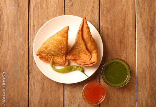 Two samosas kept on a white dish with chilly. Red & green chutney ket in a bowl.