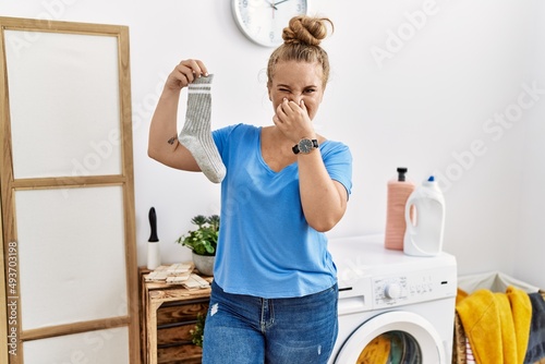 Young caucasian woman holding dirty sock at laundry room smelling something stinky and disgusting, intolerable smell, holding breath with fingers on nose. bad smell
