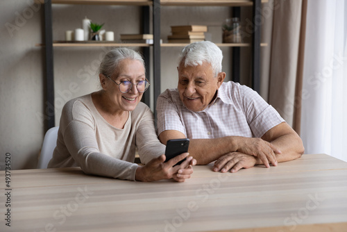 Smiling older couple sit at table staring at smart phone screen, enjoy new mobile application, lead online chat in social media with grown up children, make order, buying through e-commerce concept