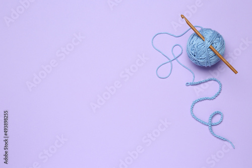 Clew of light blue knitting thread and crochet hook on violet background, top view. Space for text