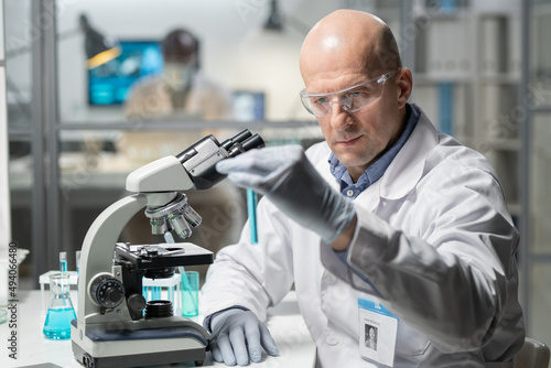 Contemporary mature male clinician in gloves, whitecoat and protective eyewear looking at blue liquid in flask while sitting in laboratory
