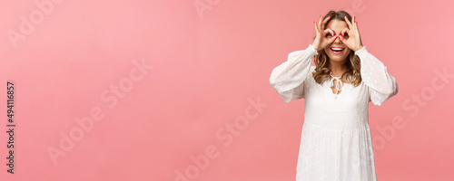 Portrait of dreamy cute and funny young blond girl seeing something interesting, looking from okay signs as making glass-mask with fingers over eyes, smiling amused, pink background