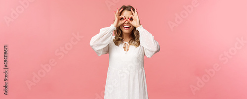 Portrait of dreamy cute and funny young blond girl seeing something interesting, looking from okay signs as making glass-mask with fingers over eyes, smiling amused, pink background