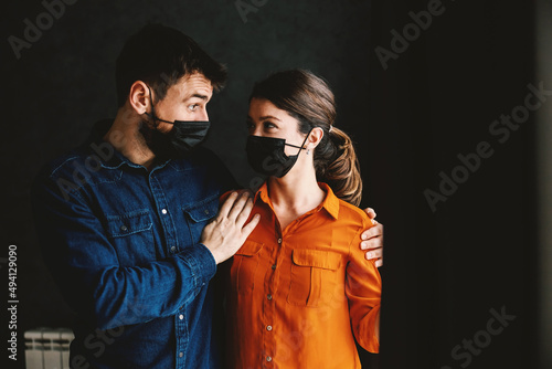 Couple with face masks standing next to window and looking at each other. 