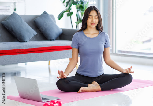 Asian young happy peaceful calm female model in casual sporty outfit sitting crossed legs in lotus position on yoga mat learning studying online meditation class via laptop computer in living room