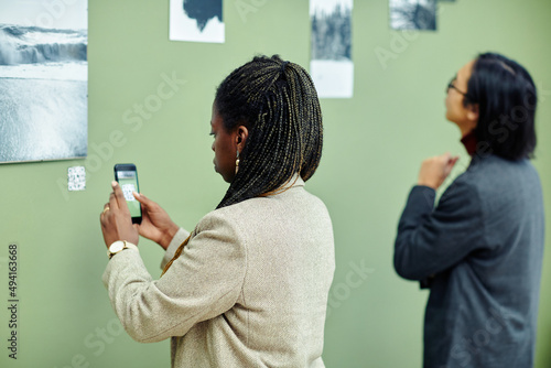 Young African American woman attending exhibition in modern art gallery scanning QR code using smartphone to find more information about photo on wall