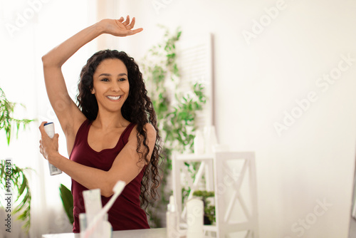 Beautiful millennial woman using spray deodorant for armpit zone, smiling at herself in mirror at bathroom, free space