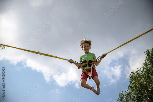 The boy is jumping on a bungee trampoline. A child with insurance and stretchable rubber bands hangs against the sky. The concept of happy childhood and games in the amusement park