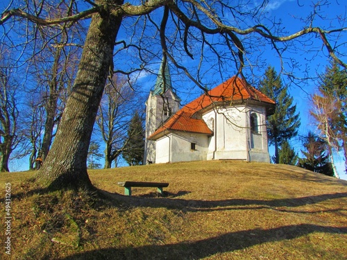 The church of St. Mohor and Fortunat at the top of Osolnik hill in Slovenia with leafless trees on the side and a meadow in front