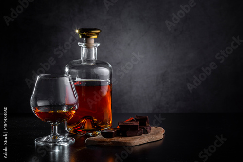 A glass of cognac, cognac in a bottle and pieces of chocolate on a dark background. Copy space