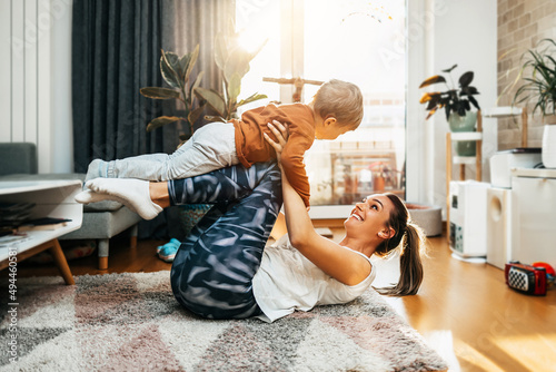 Beautiful young mother practicing fitness exercising and yoga together with her adorable little son. They are enjoying, playing and smiling in home living room.