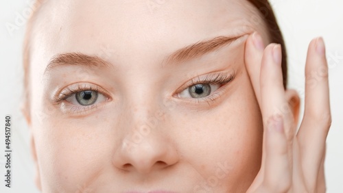 Extreme close-up shot of good-looking red haired female skin care model lifts her eyes on white background | Droopy eyelid prevention concept